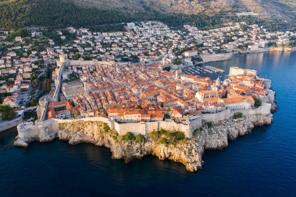 Aerial view of Dubrovnik's old town surrounded by massive stone walls and the Adriatic Sea, with a modern cityscape advocating sustainable tours in the background.