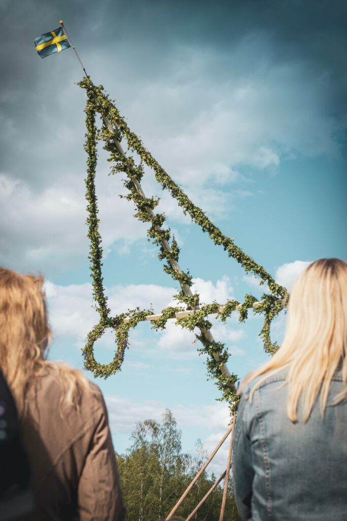 Two people seen from behind observing a traditional Swedish maypole adorned with greenery and flowers during the summer solstice, under a blue sky with a Swedish flag at the top.