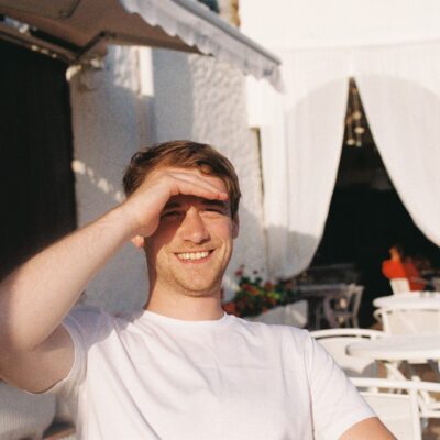 Young man shielding his eyes from the sun, smiling outdoors with a white patio and plants in the background on "The Real Word.