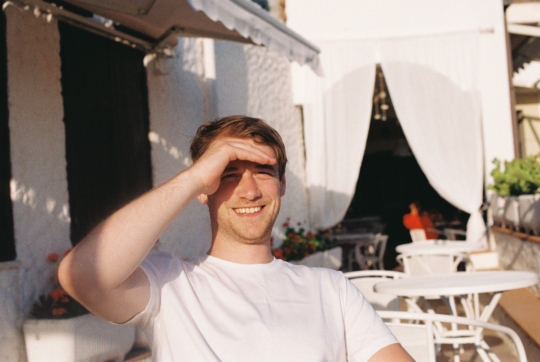 Young man shielding his eyes from the sun, smiling outdoors with a white patio and plants in the background on "The Real Word.