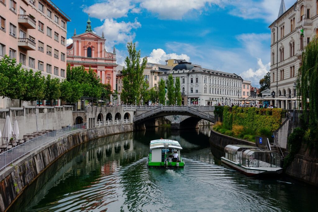 A boat cruising under a bridge on the Ljubljanica River in Ljubljana, with classical European architecture and blue sky in the background, showcasing one of the family-friendly European