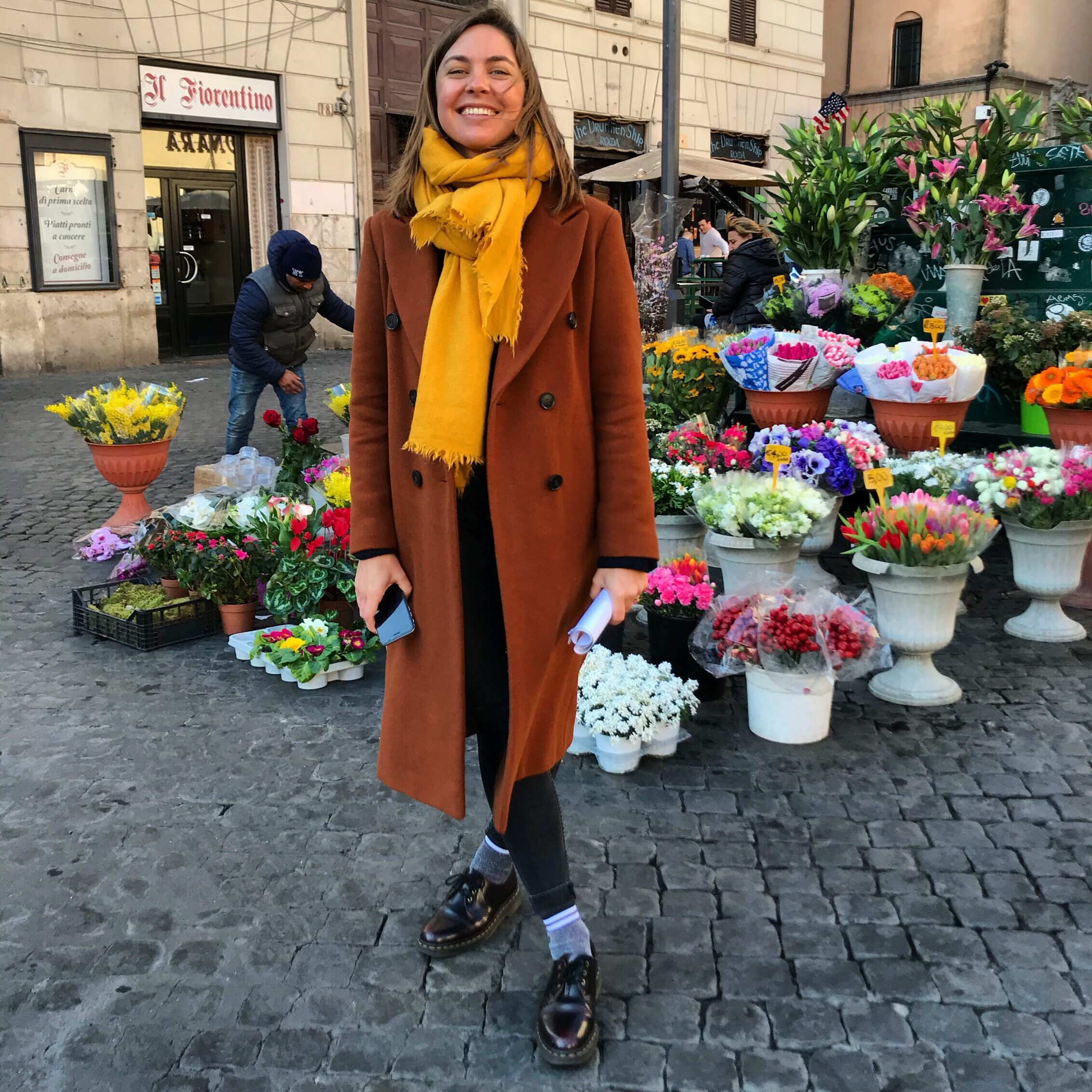 Photo of Ginny standing in front of a flower stool in a cobbled city market square