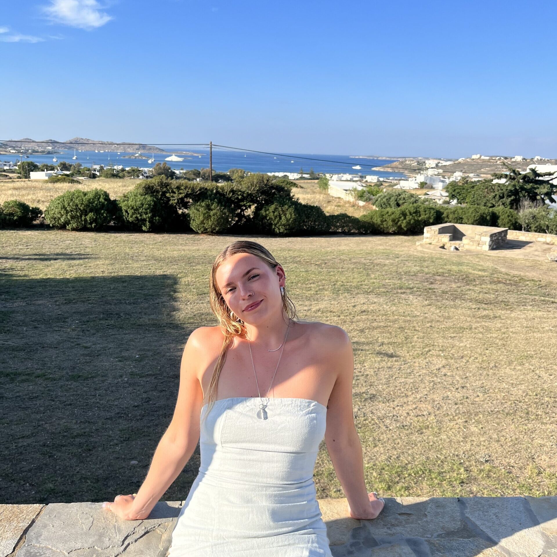 Photo of Jess sitting on a low wall, with a view of the sea and blue sky in the background