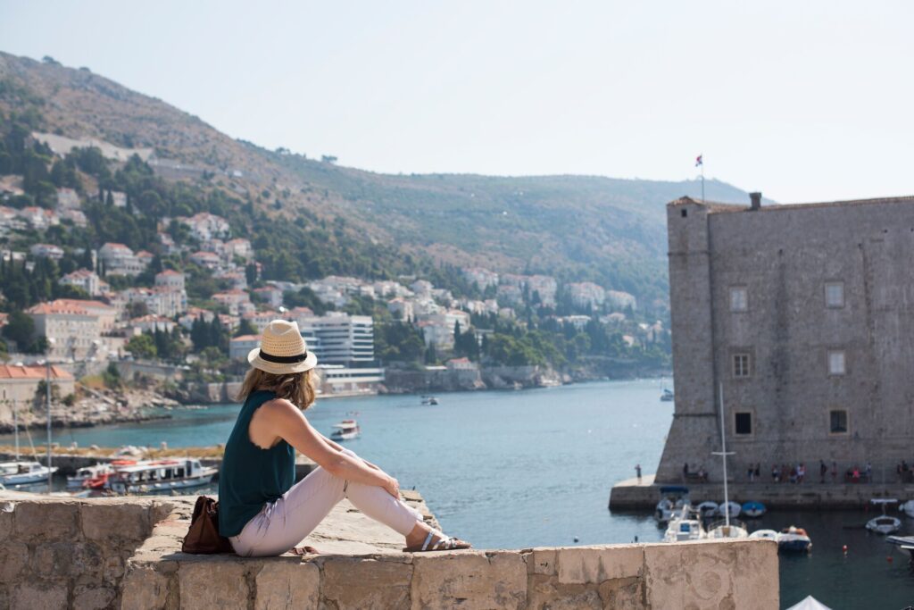 A woman in white capri pants, a teal singlet and fedora hat sitting on the wall of Dubrovnik looking out to sea
