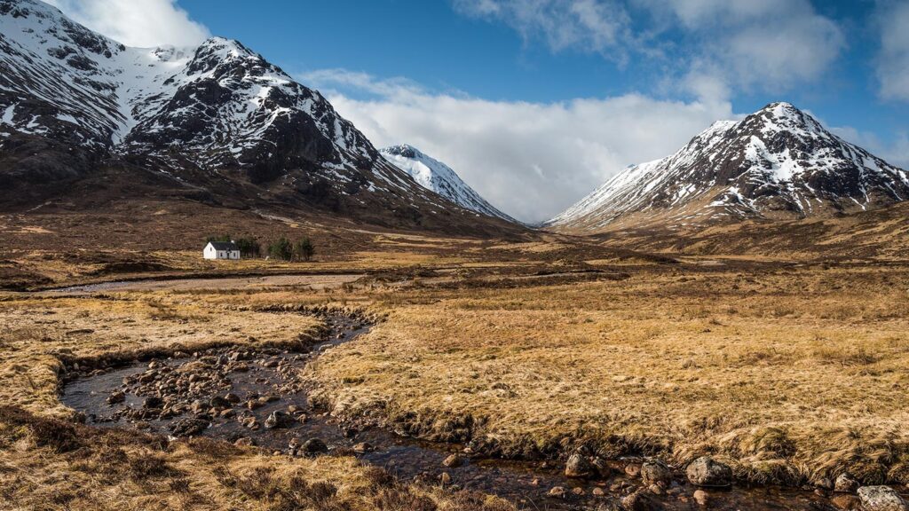 A small house in the middle of oat-colored fields with snowy peaks rising in the background in Scotland's Scottish Highlands