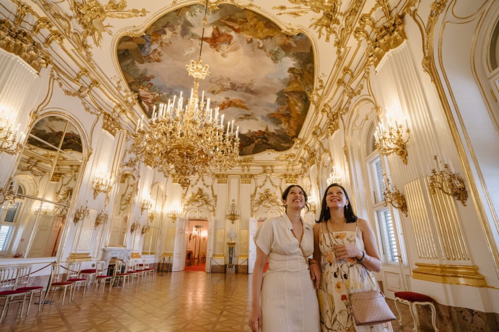 Two women walking through an extravagant gilded room with a chandelier at Schonbrunn Palace