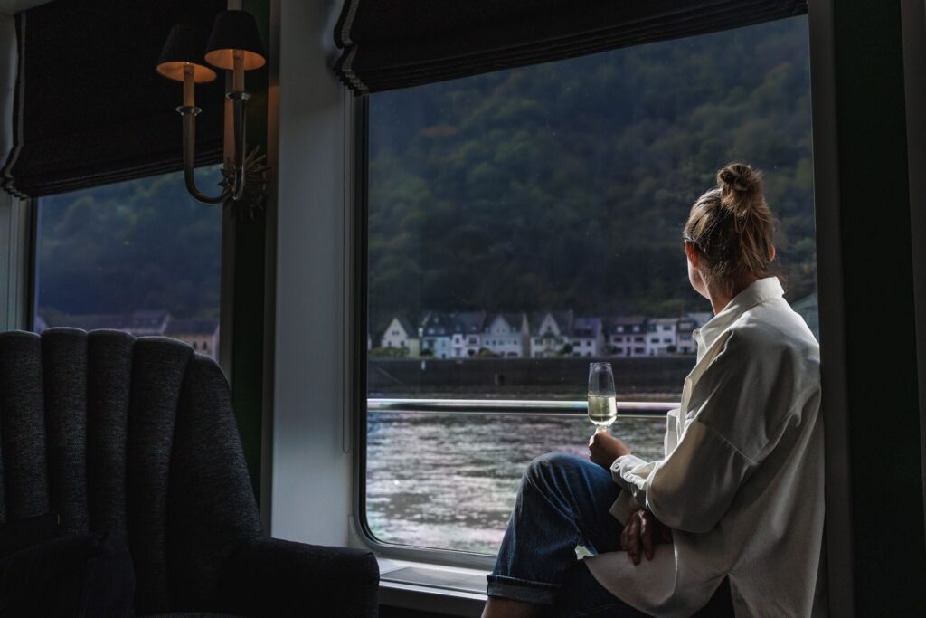 A woman with a glass on sparkling wine sitting in the window of a river cruise boat looking at traditional houses on the shore.