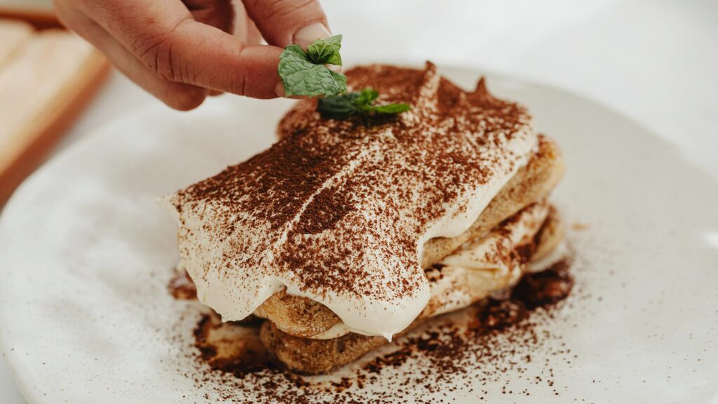 A hand placing a mint leaf on top of a rustic tiramisu in a restaurant