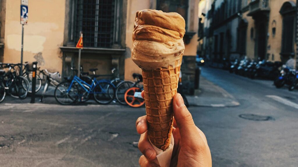A hand holding a cone of gelato with two scoops on a street in Italy