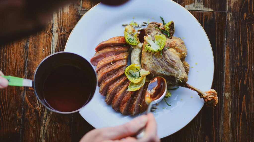 A chef spooning gravy over a dish of Confit de Canard duck on mashed potato