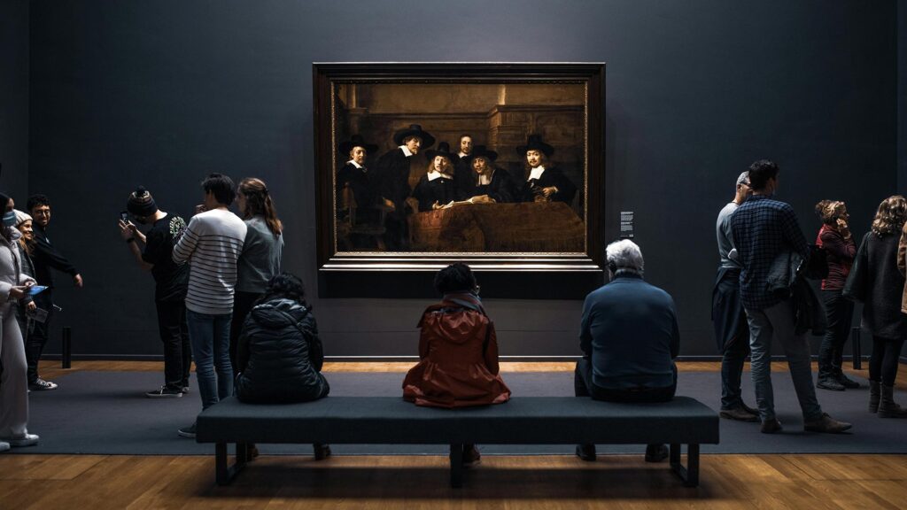 A dark room with a dozen or so people viewing a Dutch masterpiece at the Rijksmuseum