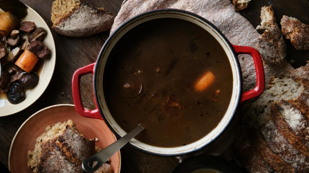 A red pot on the table filled with rich Boeuf Bourguignon stew with a crusty loaf of bread ot one side