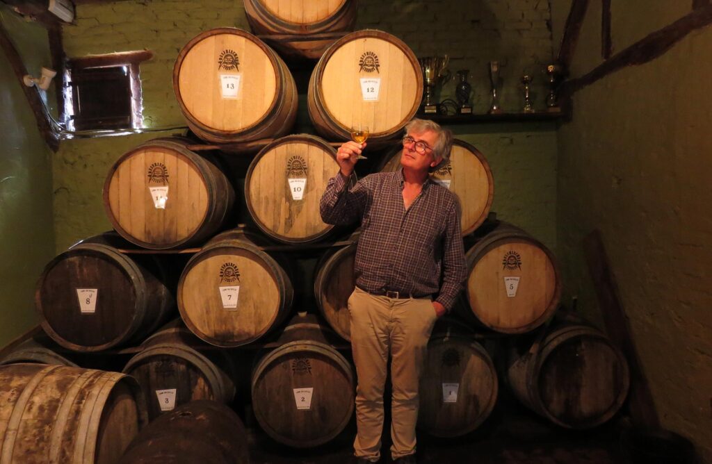 Michel from La Ferme Des Ruelles holding up a glass of cider while standing in front of a stack of barrels ageing local cider