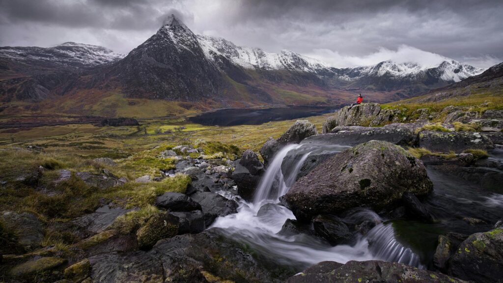 Man sitting on the edge of a rocky waterfall overlooking a valley with a large snow-capped mountain in the distance.