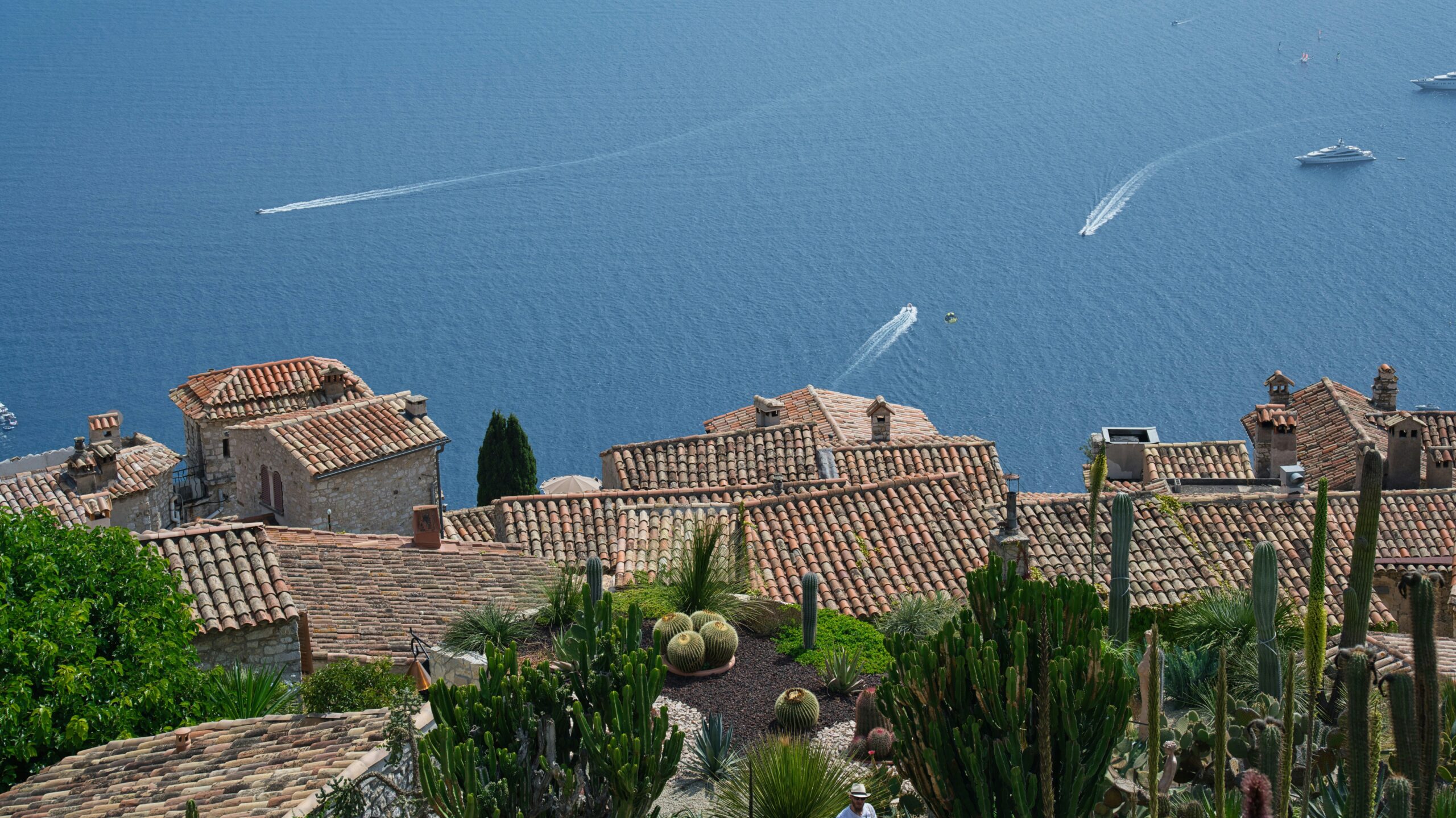 View of terracotta tiled rooftops in front of a blue sea with boats streaking the water