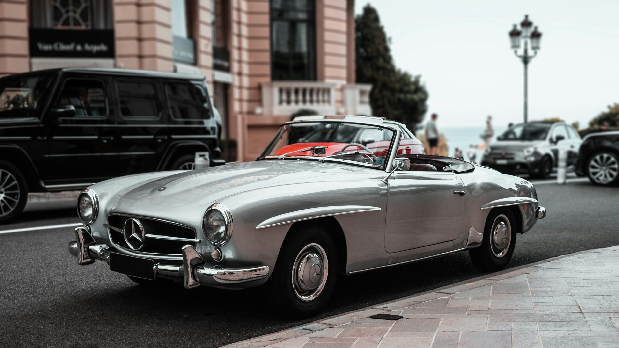 Vintage silver convertible parked on a street beside a luxury hotel, with the sea visible in the background.