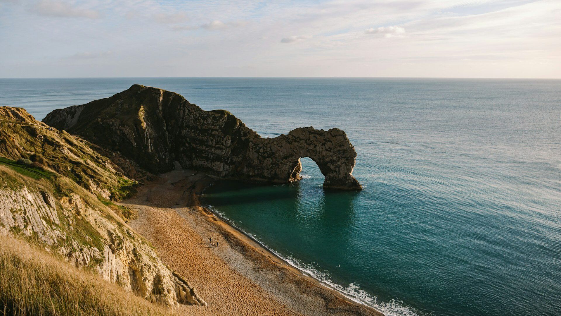 Cliff-top view of Durdle Door — a limestone sea arch, beyond a sun-dappled beach