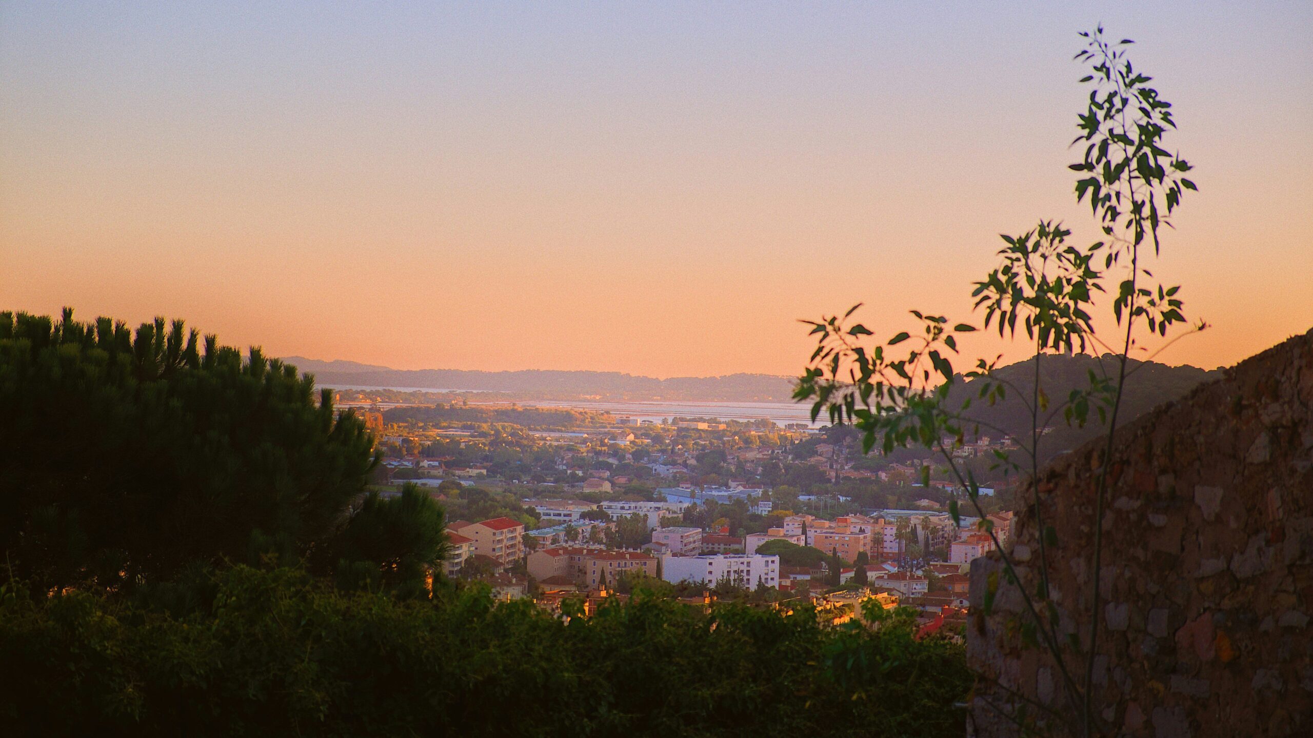 Sunset view across a coastal town in southern France, framed by trees and rolling hills.