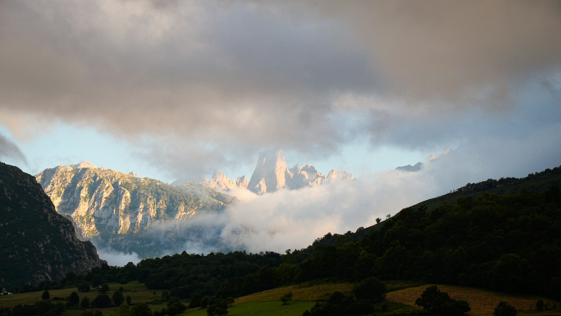 View of sunlit mountains appearing through clouds beyond a dark hillside