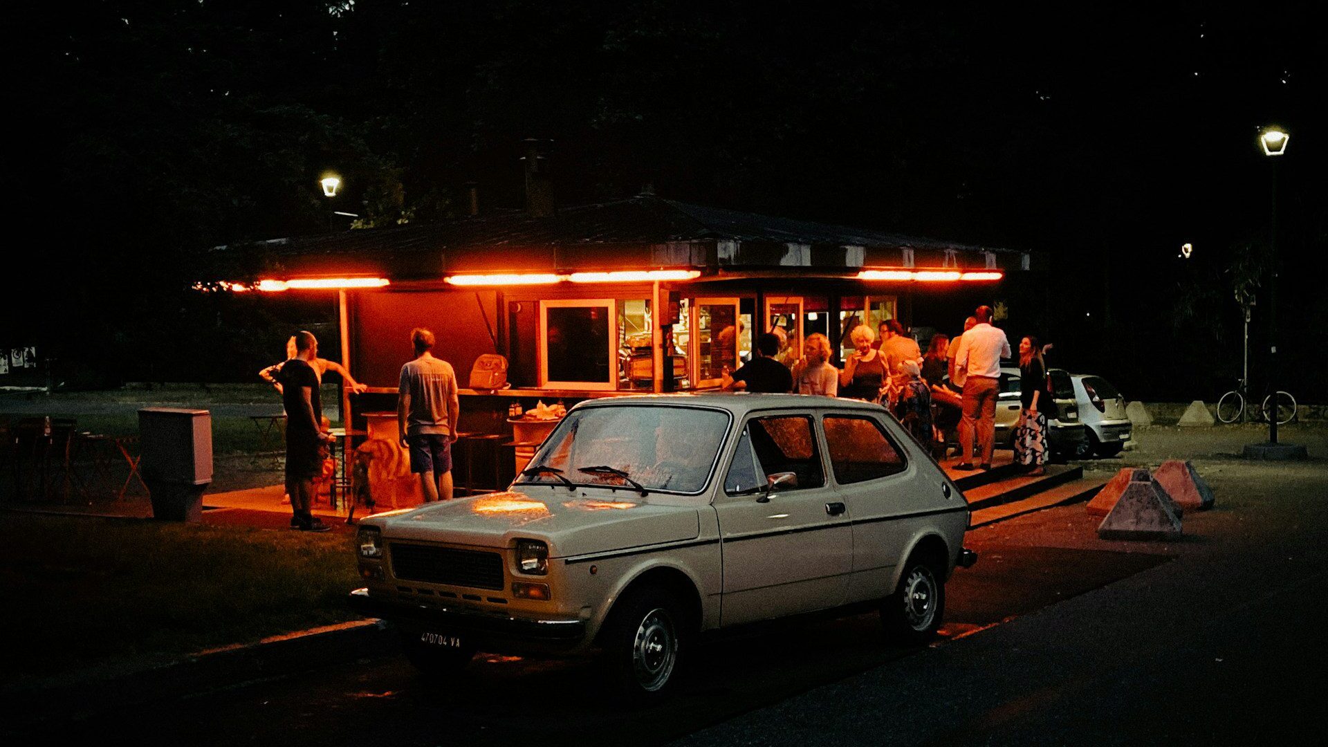 People chatting and drinking under red lights at an outdoor bar, with a vintage Fiat car in the foreground.