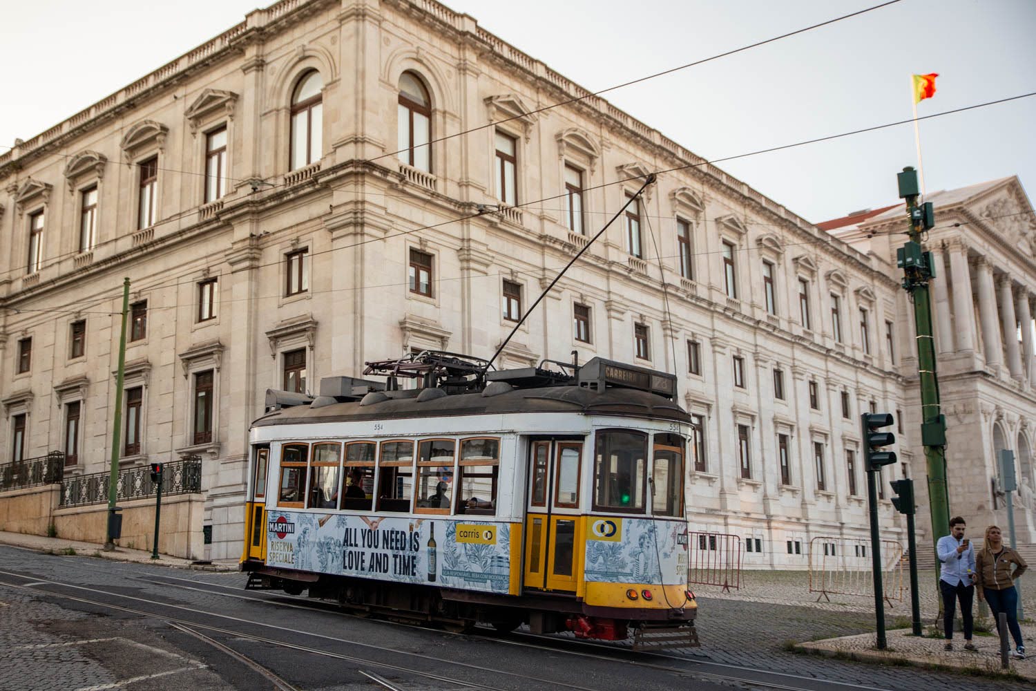 Yellow tram heading down a hilly street in front of a grand ornate building in Lisbon