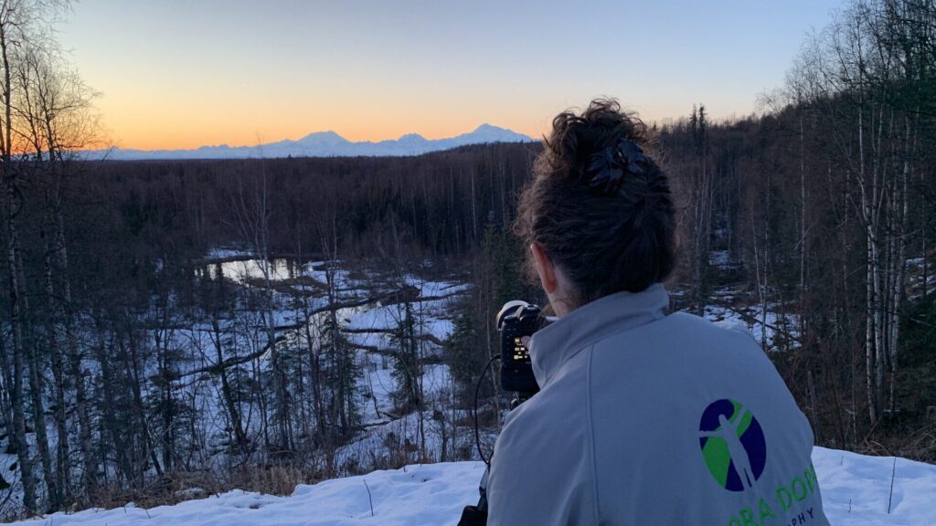 A photo of Dora from behind as she looks through her camera towards a mountain range with the sun setting behind it.