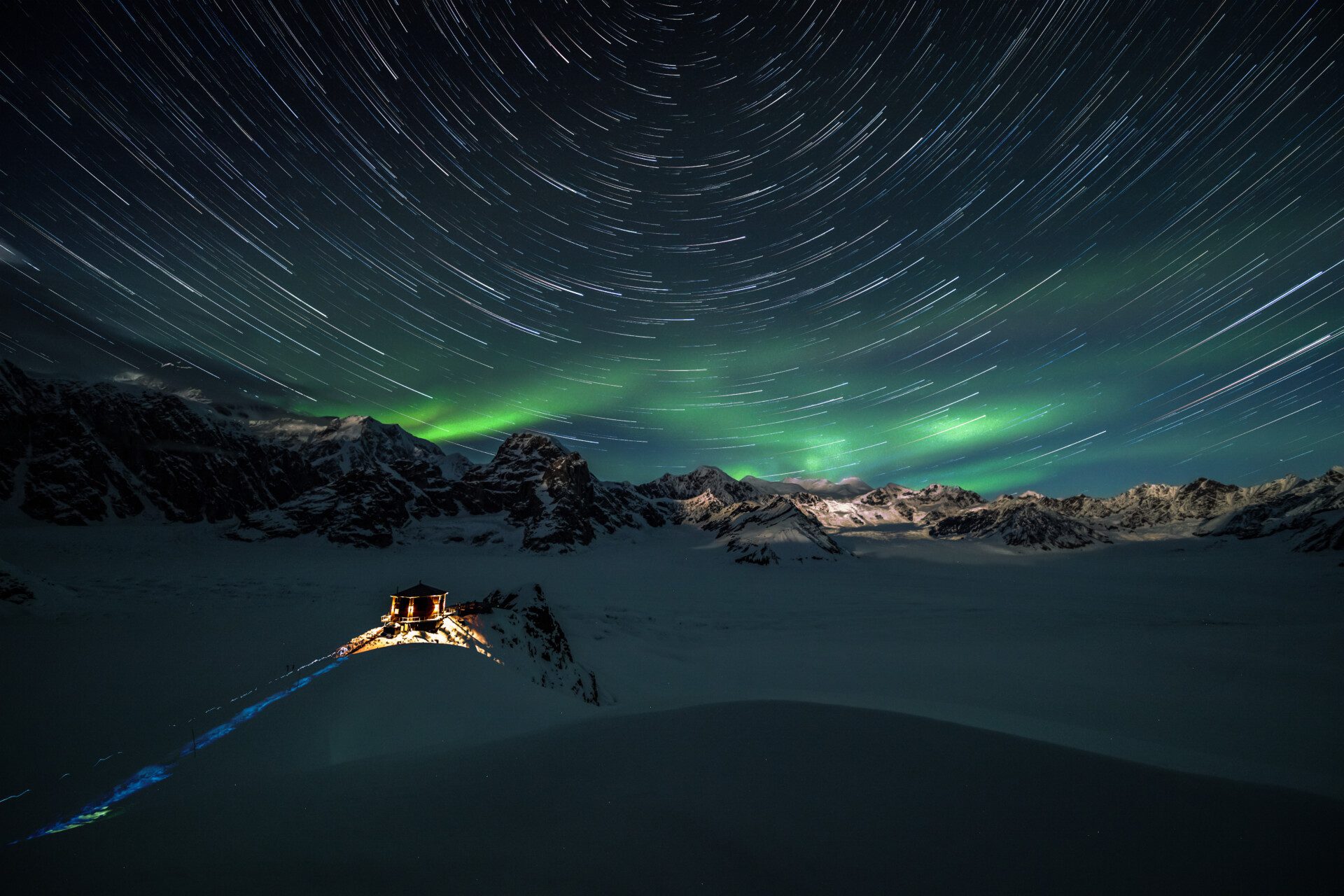 A long-exposure of stars in the night sky with northern lights visible on the mountainous horizon