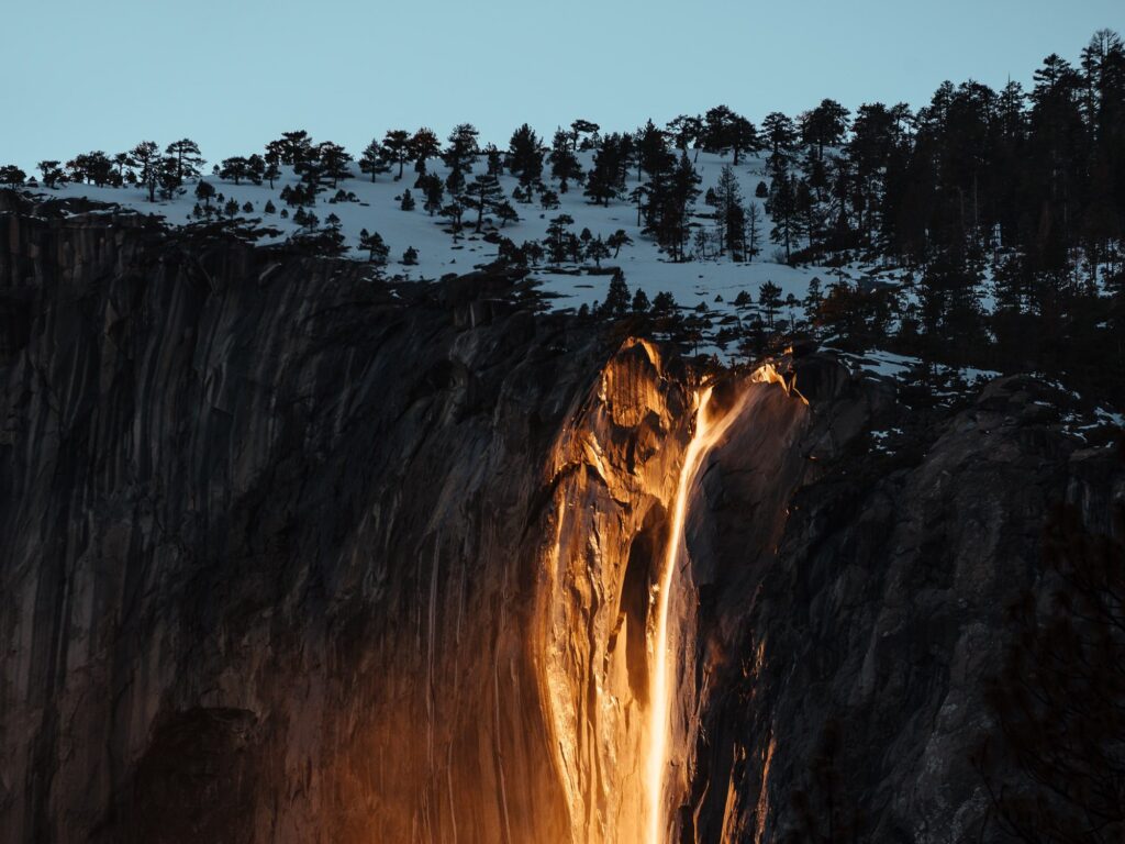 Firefall effect on Horsetail Fall in Yosemite National Park, one of the natural wonders, with the waterfall glowing orange due to the sunset light against a shadowed rocky cliff.