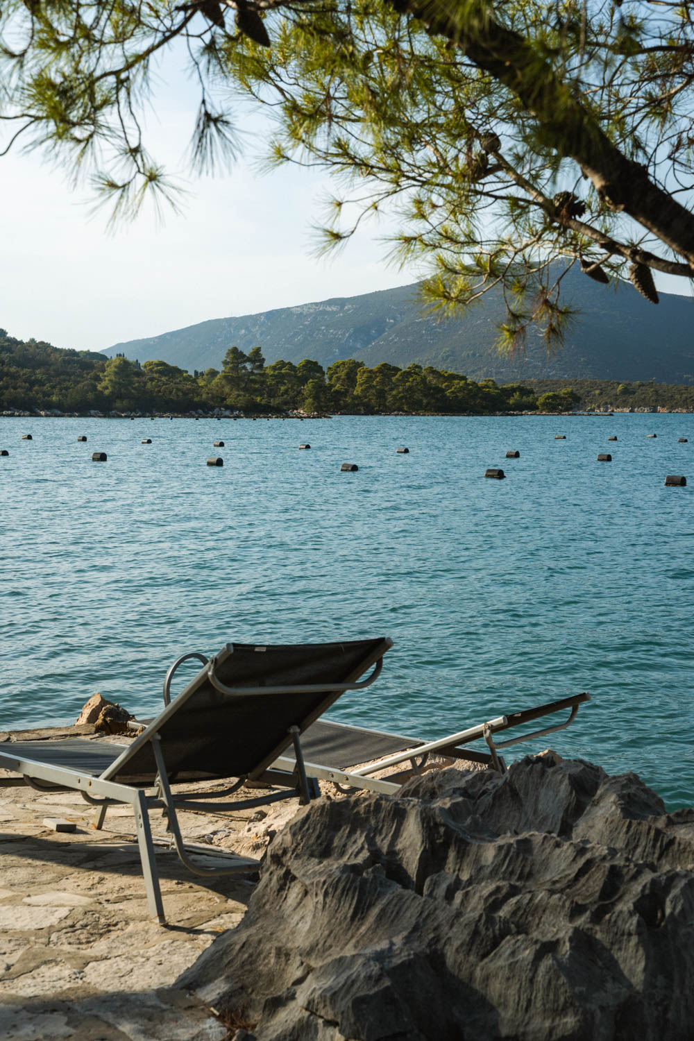 Two sun loungers sitting on a paved area by the edge of the Adriatic Sea, with islands and mountains in the background.