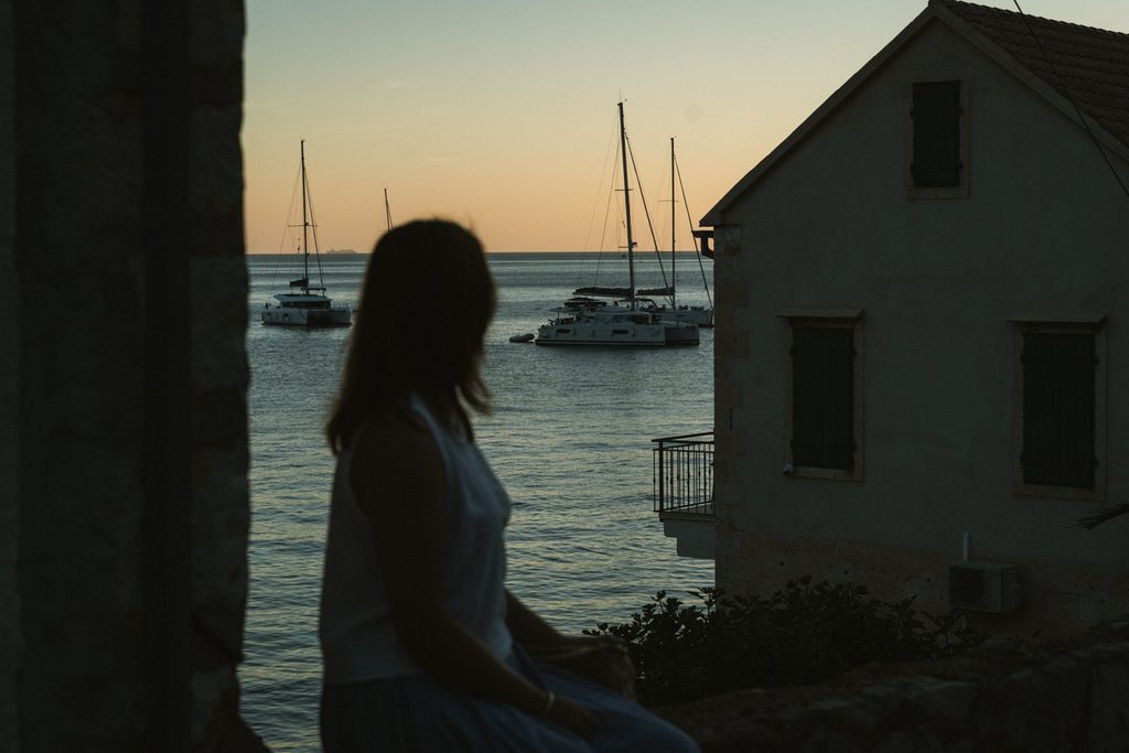 Silhouette of a woman sitting on a stone wall looking out towards sail boats on the sea at sunset.