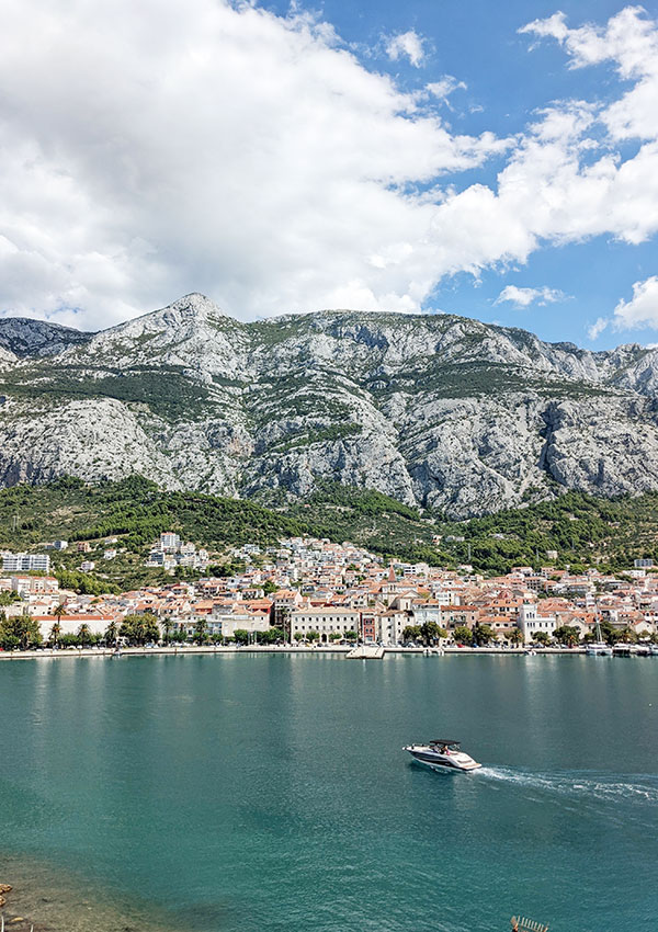 A boat speeds across a blue sea towards a coastal town.