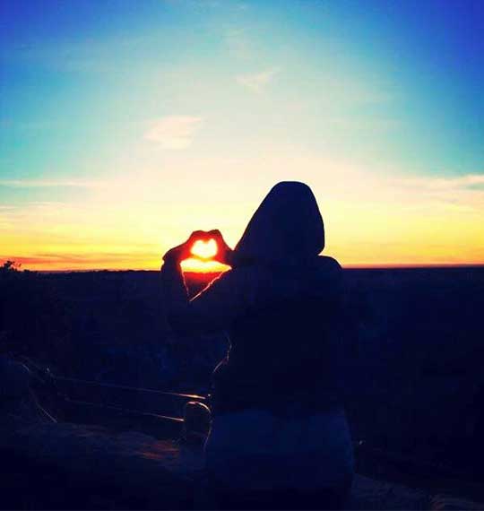 Capturing travel moments at sunset, a person forms a heart with her hands.