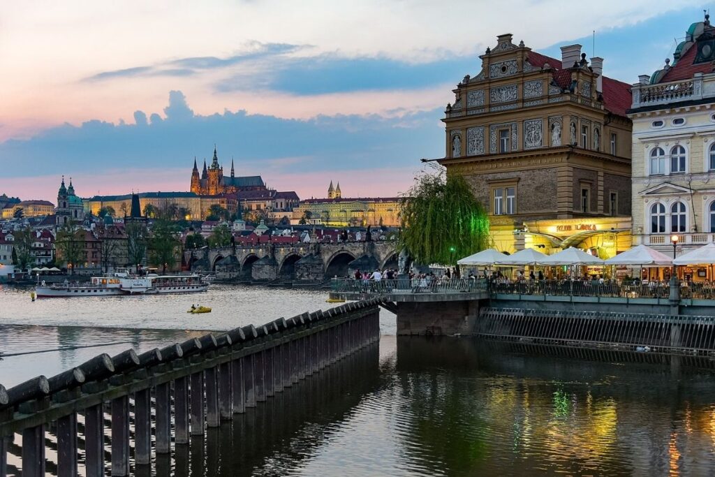 River in the forefront with buildings in the background, umbrellas and on-street dining is before the buildings.
