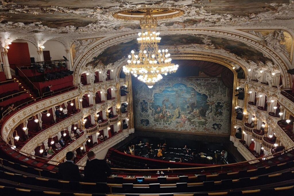 Inside Prague State Opera House, red velvet seats and ornate ceiling and chandelier in the middle. Stage down below, with room for an orchestra in front.