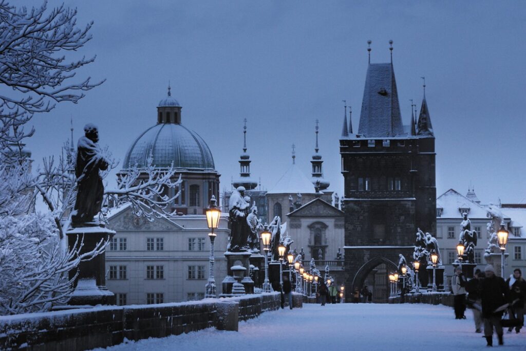 Winter time, Charles Bridge in Prague. The bridge has a layer of snow, with statues either side snowy too. Buildings in the background.