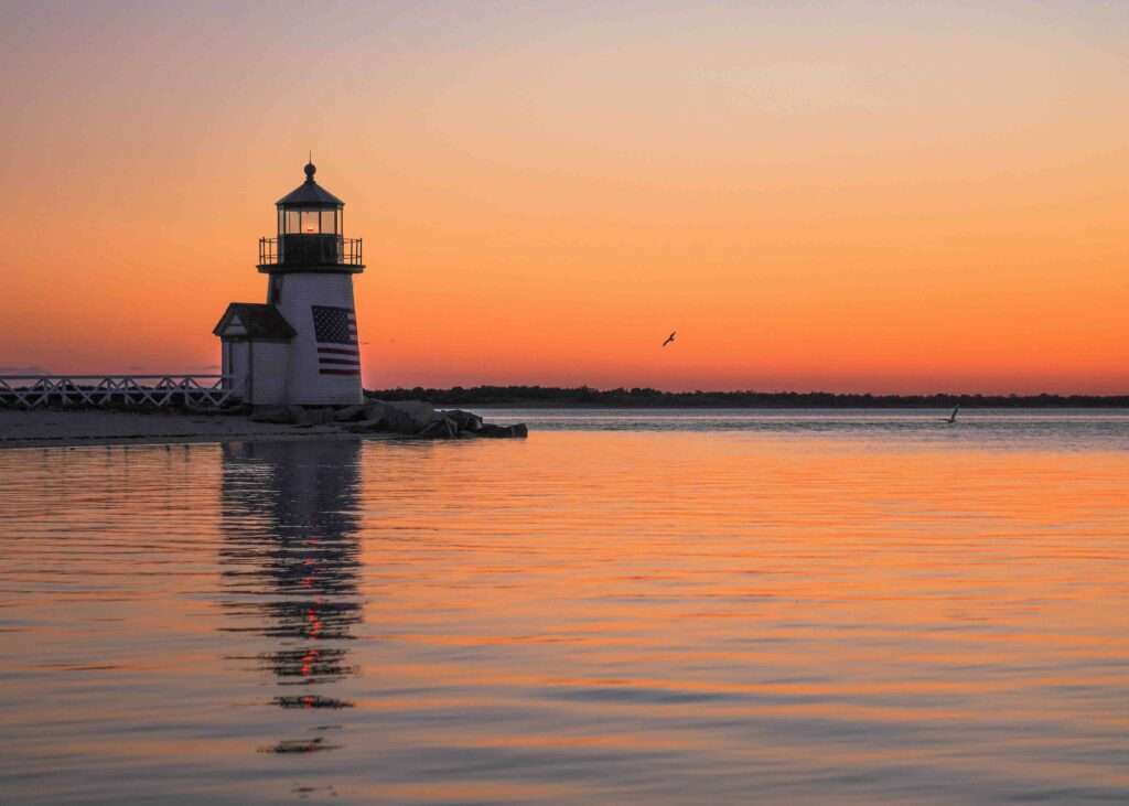 Sunset in Nantucket, unique family Christmas vacation ideas. Lighhouse to the left of the image with a large US flag on the outside of it.