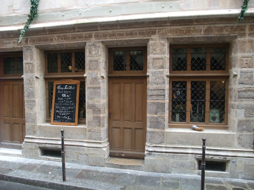 Stone house from 1407 in Paris, brown door in the centre, with windows either side of the door.