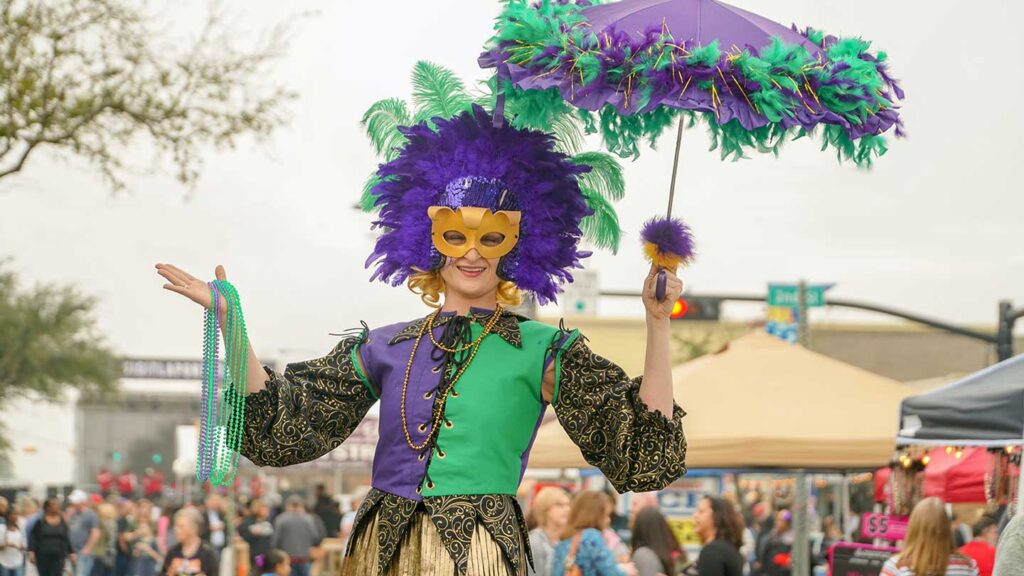 A woman in a vibrant mardi gras costume with a purple feather headdress and mask, holding beads, parades at one of the bucket list events, a busy outdoor festival.