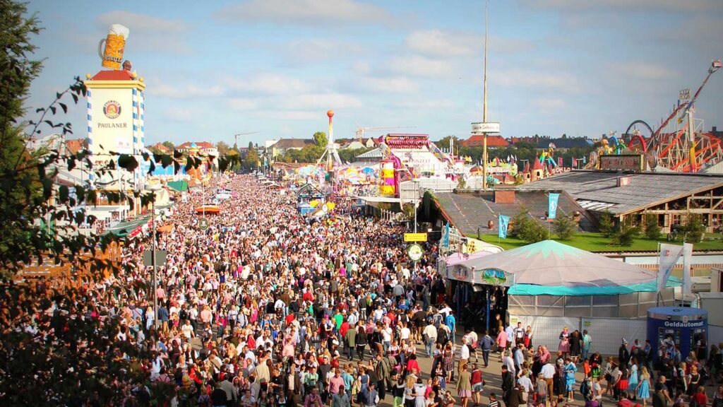Crowded bucket list outdoor fair with numerous visitors walking between amusement rides and festival booths under a clear sky.