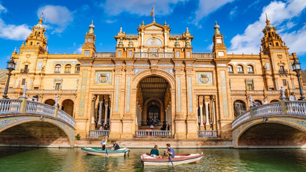 people rowing boats in front of grand building seville spain