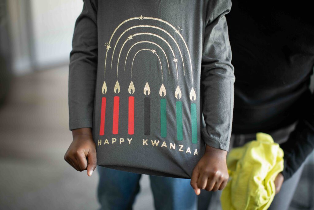 Child wearing a dark grey long-sleeve t-shirt. The child is pulling it down to show what is on the shirt. It says ‘Happy Kwanzaa’ with the seven candles above the writing.