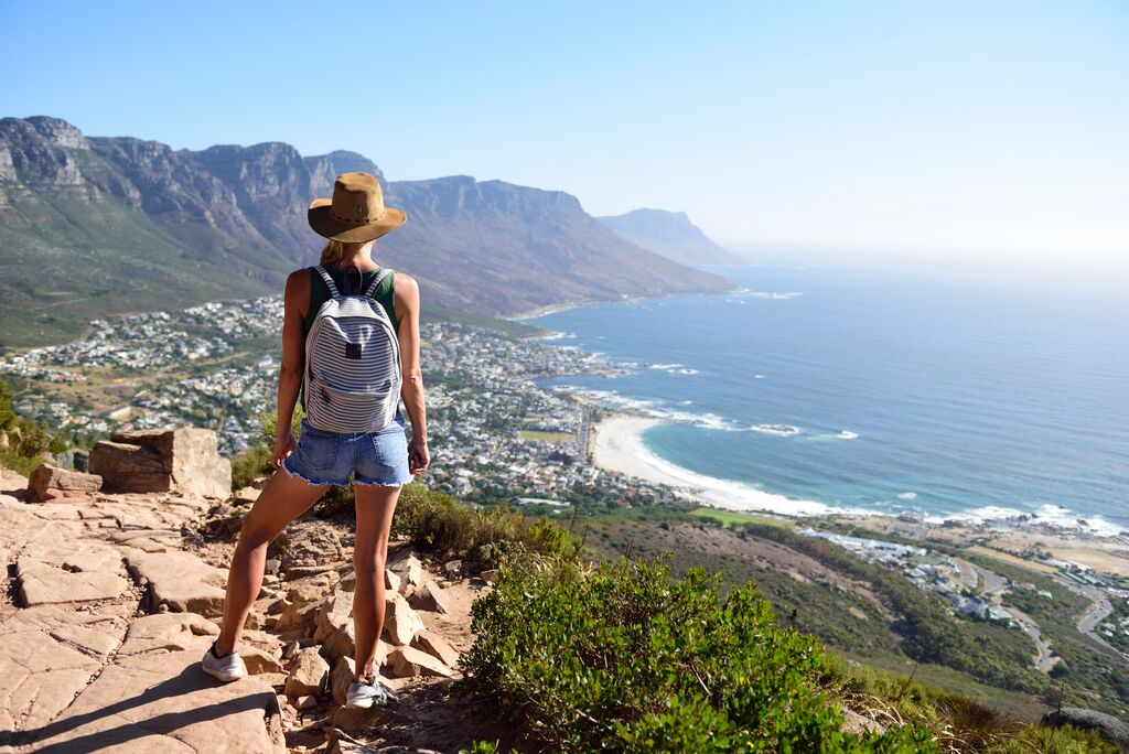 Woman in a hat and backpack standing on a rocky overlook, admiring a coastal view with mountains in the distance during her spring tour.