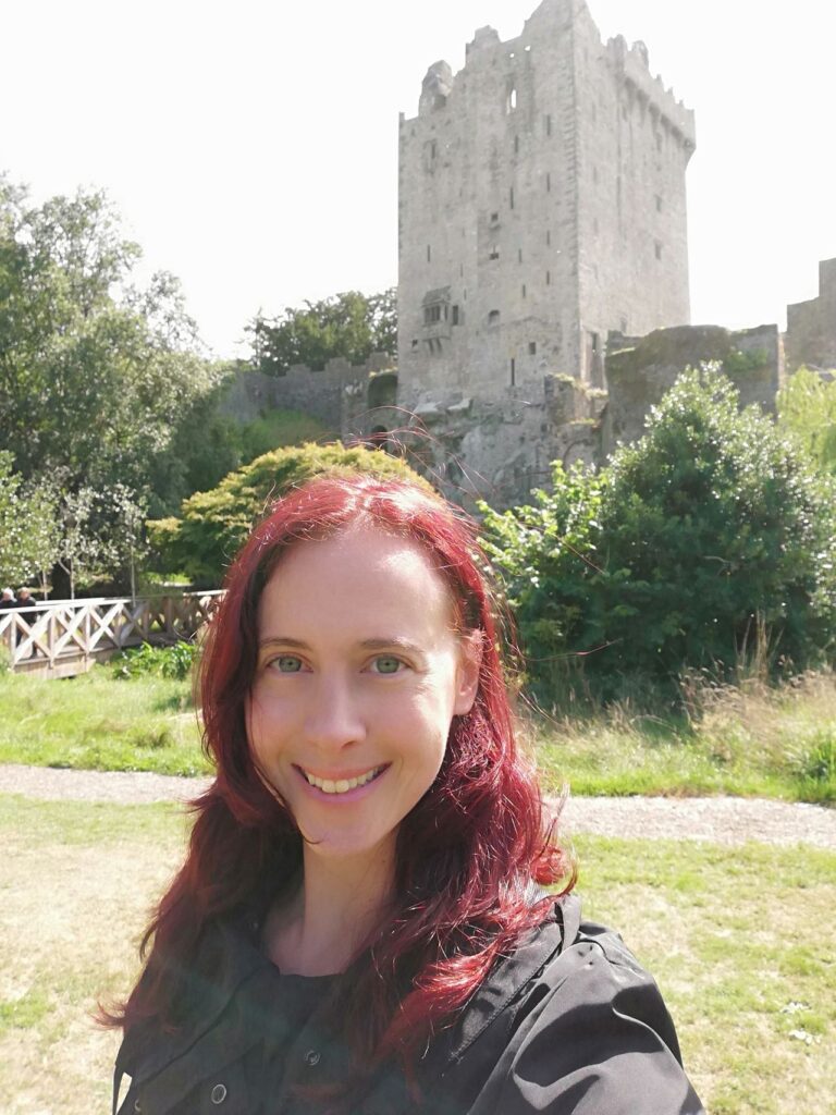 A girl with red hair and green eyes smiles for the camera in Ireland in front of an old ruined tower