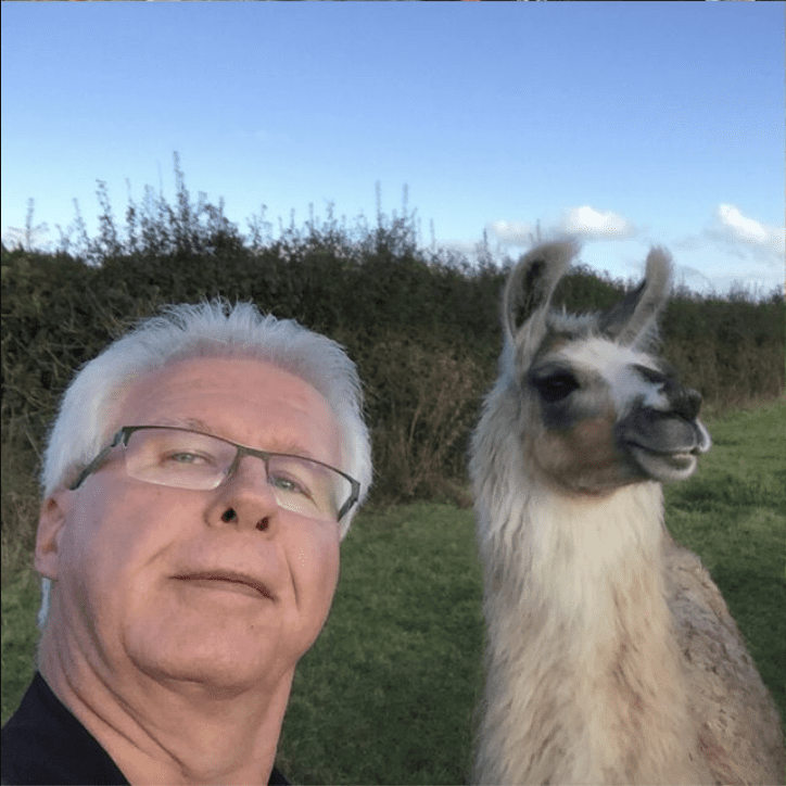 A man with white hair and glasses poses for a selfie with a llama in the background