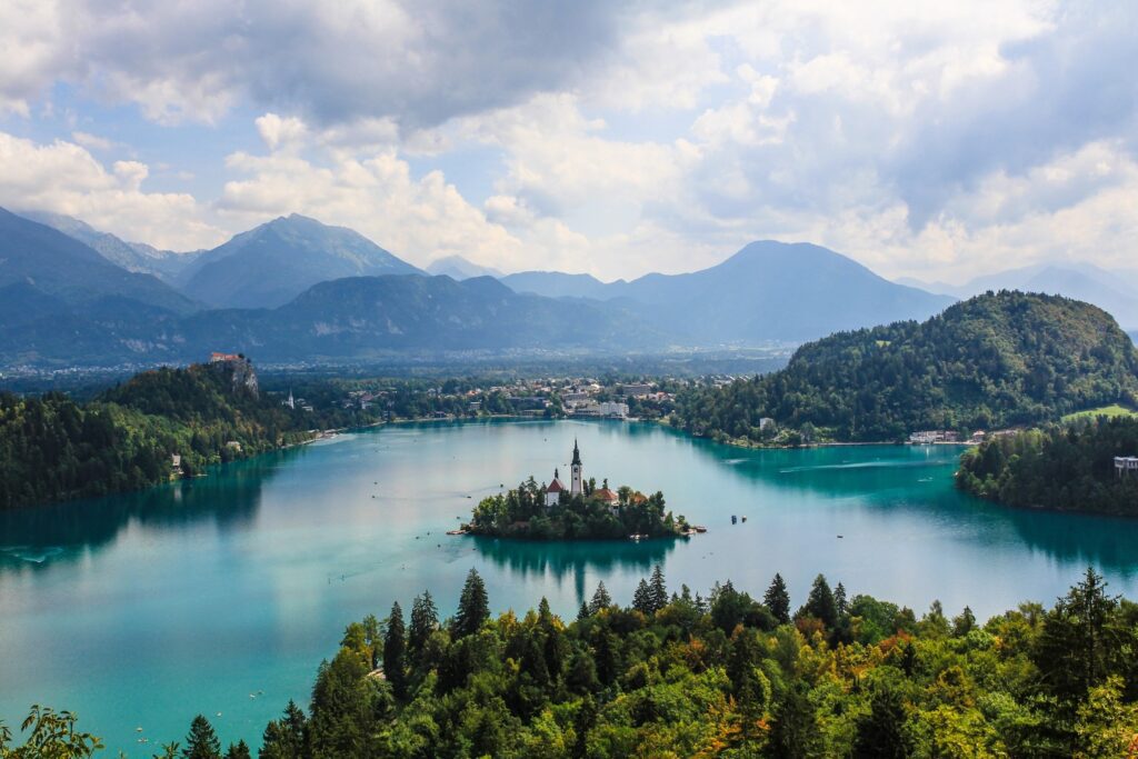 Panoramic view of Lake Bled in Central Europe, featuring a small island with a church, surrounded by lush greenery and mountain backdrop under a cloudy sky.