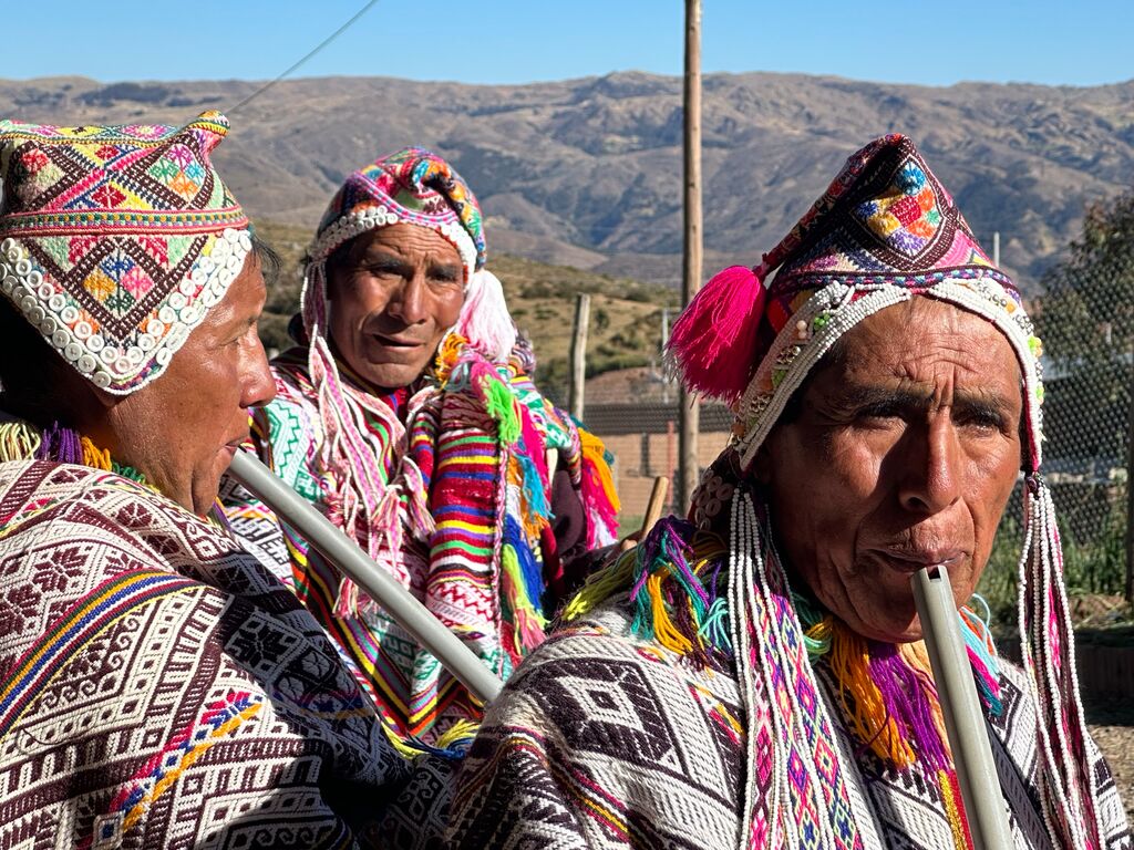 Three Peruvian men in traditional dress stand with the Andes in the background, two of them playing  wind instruments