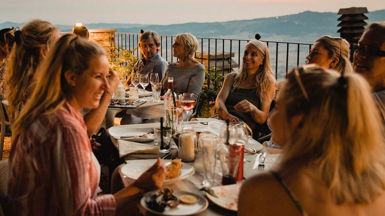 A group of male and female diners sitting around a table sharing food, wine and conversation. View of distant hills in background