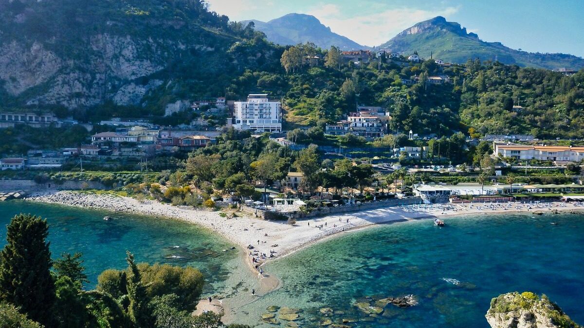 View of a beach with turquoise water, buildings in the middle distance and hills in the far distance
