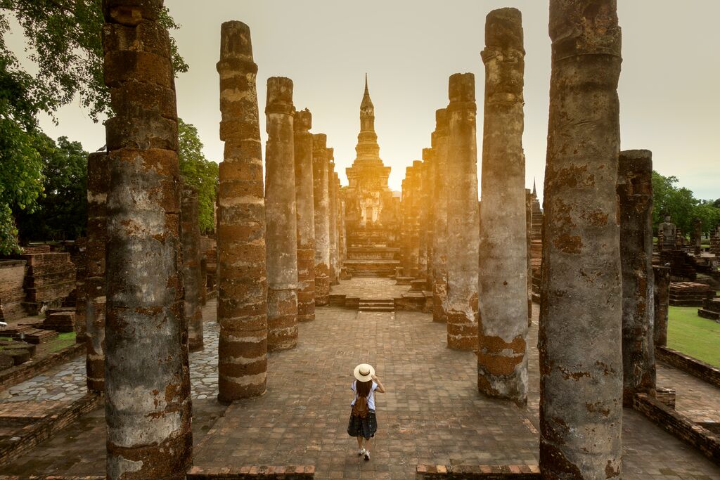 A woman with a straw hat walks away from camera, toward a Thai temple while huge pillars line the sides of the walkway.