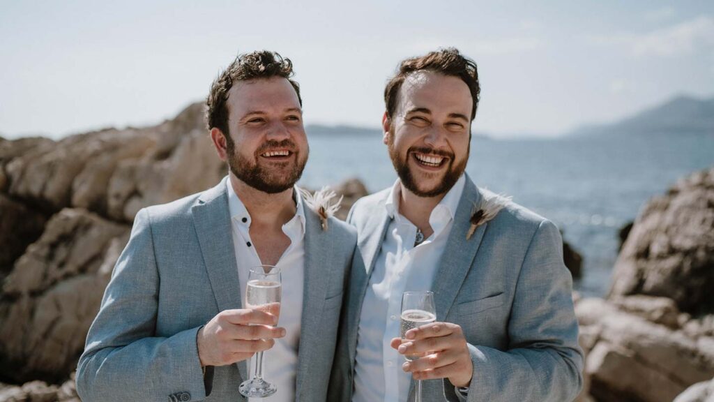 Craig and Graham pose for a photo in their grey wedding suits on a beach in Croatia
